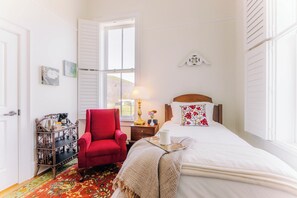 A bedroom with a bed, a red armchair, a wooden side table, a window with shutters, and a view of the outdoors. - Cozy Historic Inn Room by New River Trail (Max Meadows)