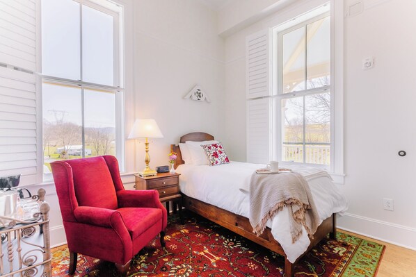 A bedroom with a bed, a red chair, a nightstand, a lamp, and a window with a view. - Cozy Historic Inn Room by New River Trail (Max Meadows)