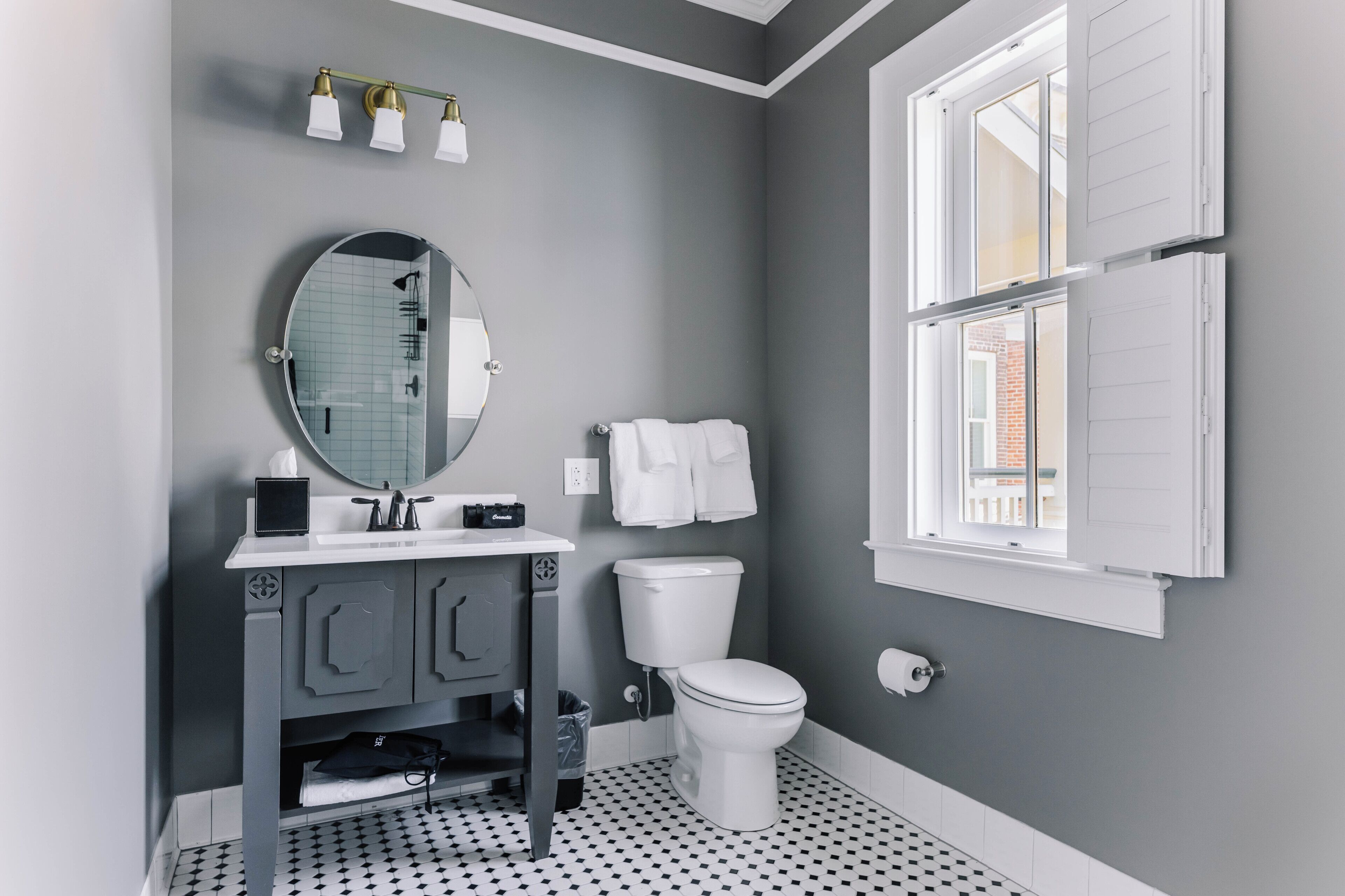A bathroom with a toilet, sink, and mirror. There is a window with shutters and a towel rack.