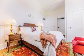 A neatly arranged bedroom with a bed, bedside lamps, a bedside table, a red chair, and a door. - First-Floor Historic Queen Room with Porch Access (Max Meadows)