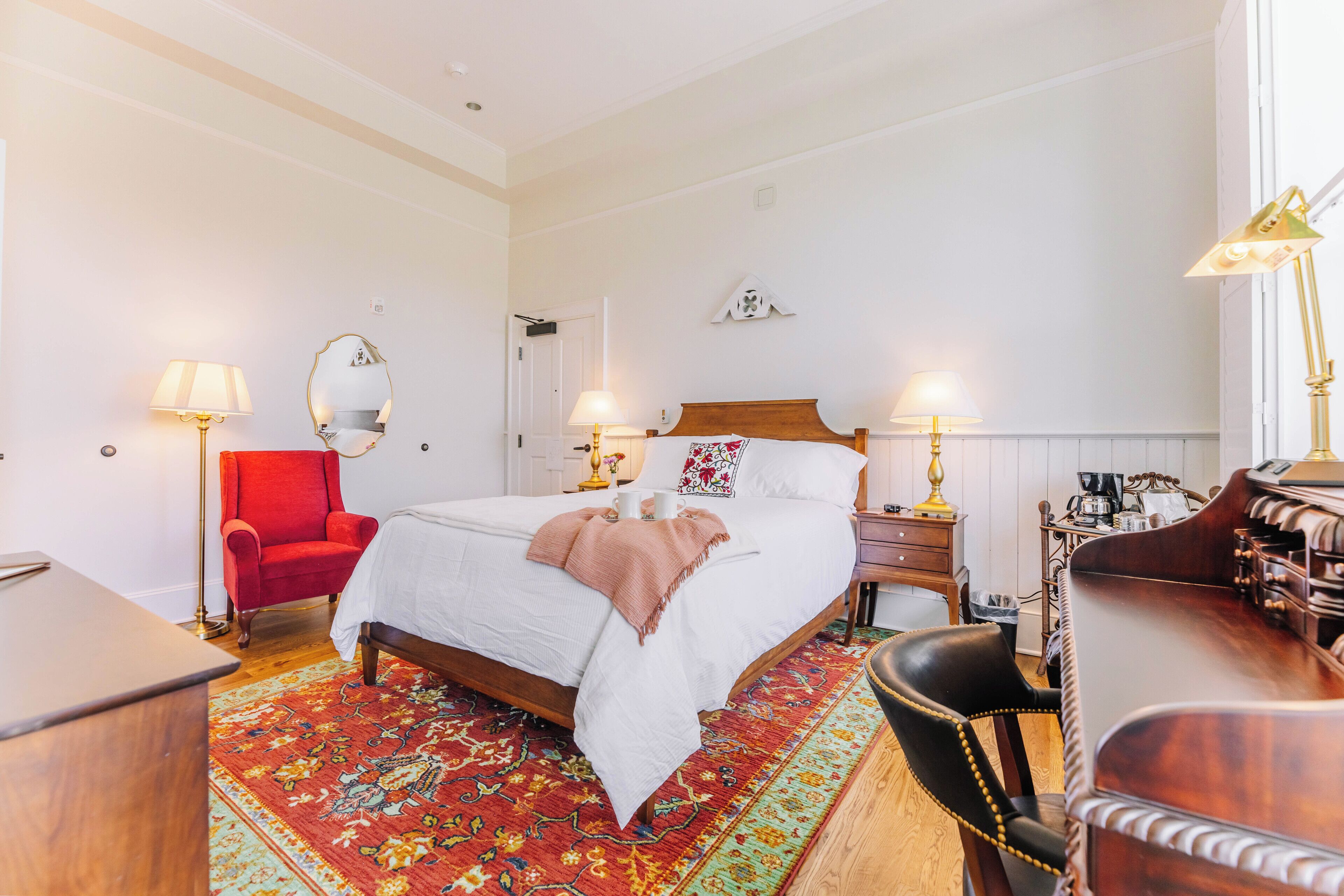 A hotel room with a bed, two bedside tables, a desk, a chair, a red armchair, and a patterned rug.