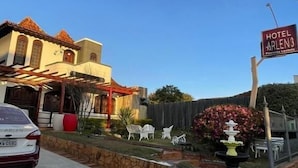 Um hotel chamado 'Hotel Arlen3' com céu claro, um carro estacionado e um jardim bem cuidado com uma fonte.