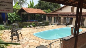 Uma área de piscina com piso de pedra, mesas e cadeiras de metal, cercada por um muro de tijolos e um portão de metal.