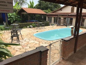 Zona de piscina con suelo de piedra, mesas y sillas de metal, rodeada por un muro de ladrillo y una reja metálica.