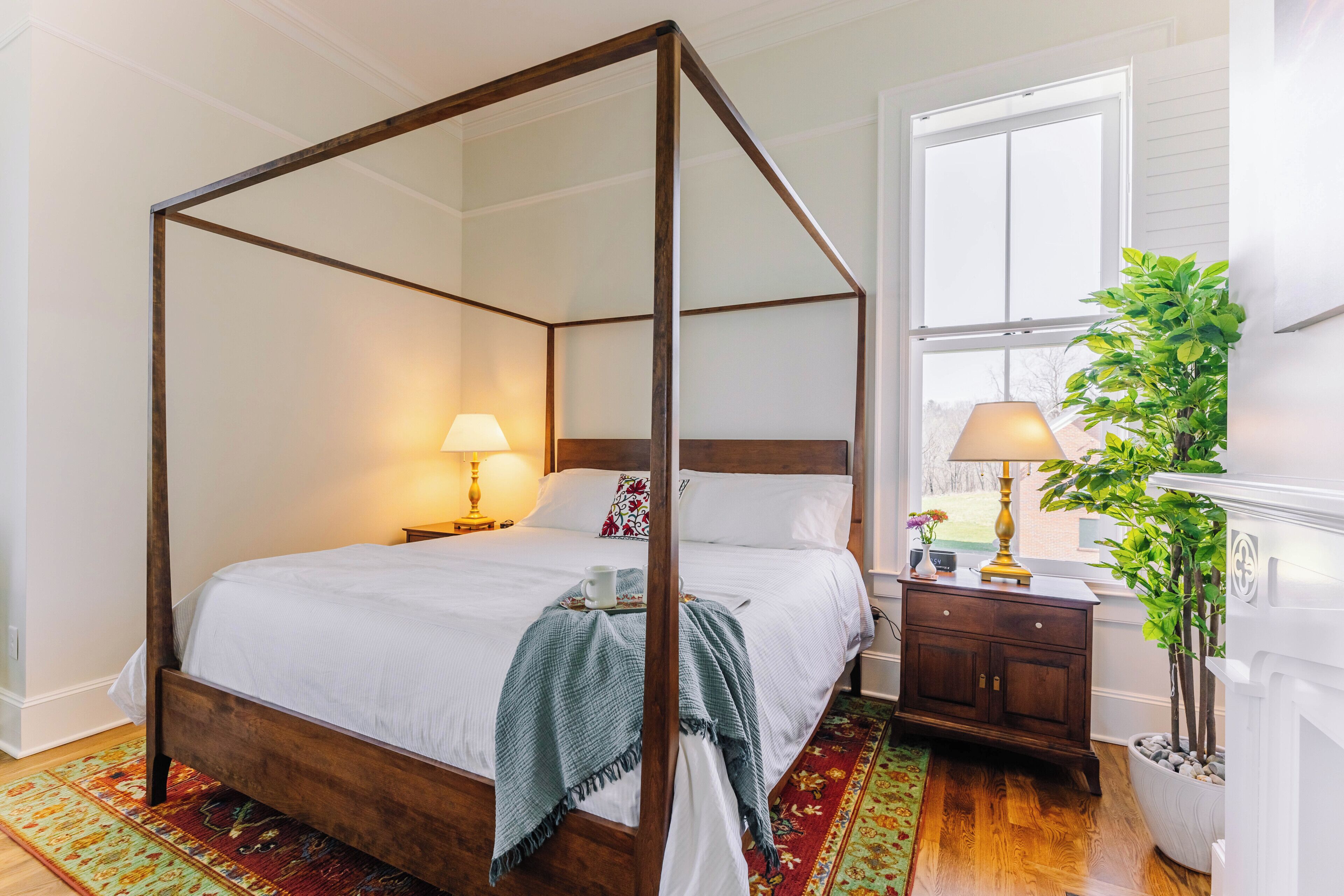 A four-poster bed with a wooden frame, a nightstand with a lamp, a potted plant, and a window with a view of the outdoors.
