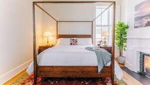 Uma cama com dossel e estrutura de madeira, roupa de cama branca e manta estampada. Há uma mesa de cabeceira com abajur, planta em vaso e um quadro na parede.