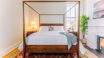 Ein Bett mit Himmel und Holzrahmen, weiße Bettwäsche und eine gemusterte Decke. Ein Nachttisch mit Lampe, einer Topfpflanze und einem Gemälde an der Wand.