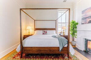 A four-poster bed with a wooden frame, white bedding, and a patterned throw. There is a nightstand with a lamp, a potted plant, and a painting on the wall. - Historic Honeymoon King Suite by New River Trail (Max Meadows)
