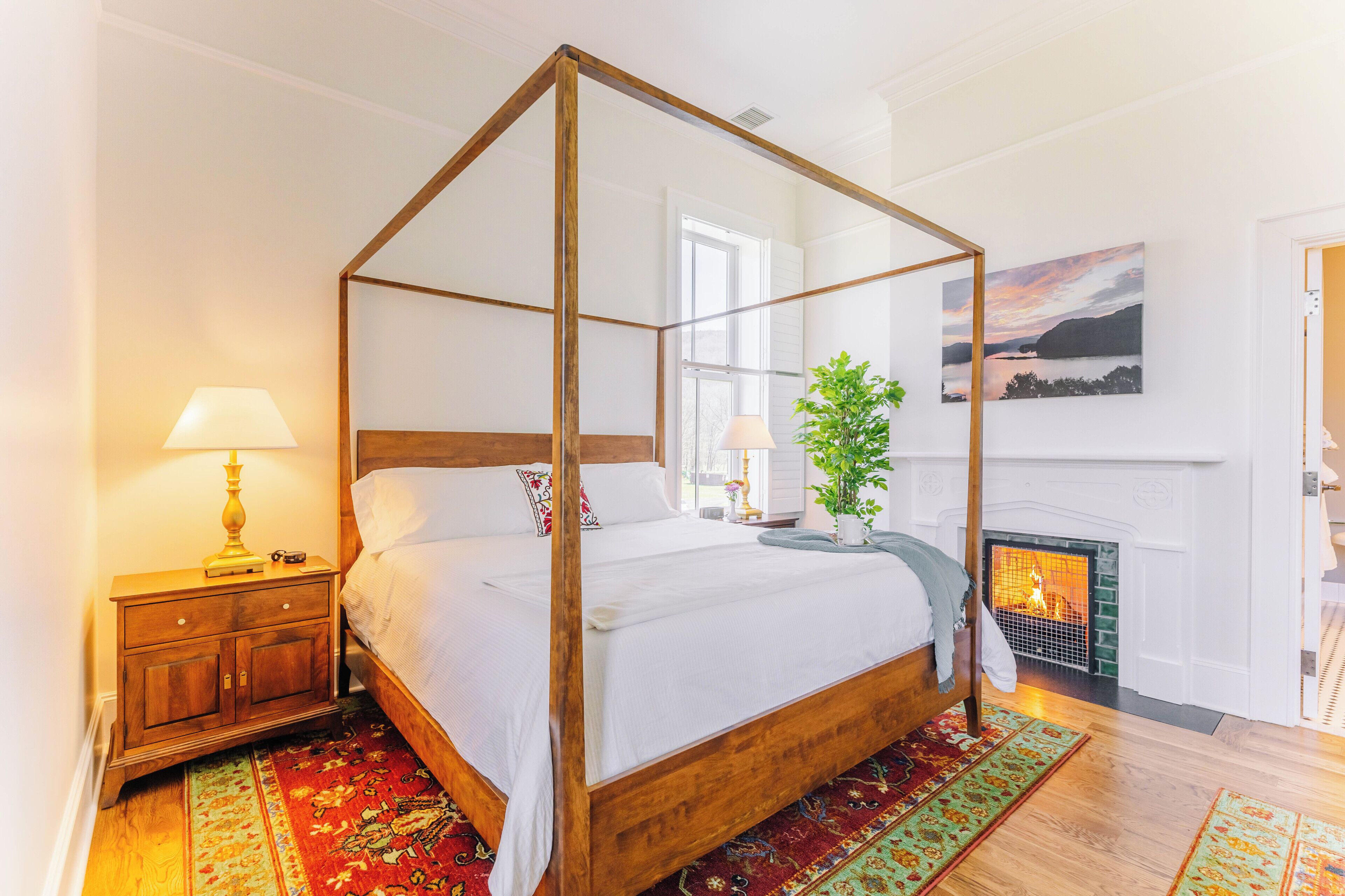 A four-poster bed with a wooden frame, a nightstand with a lamp, a fireplace, and a painting on the wall.