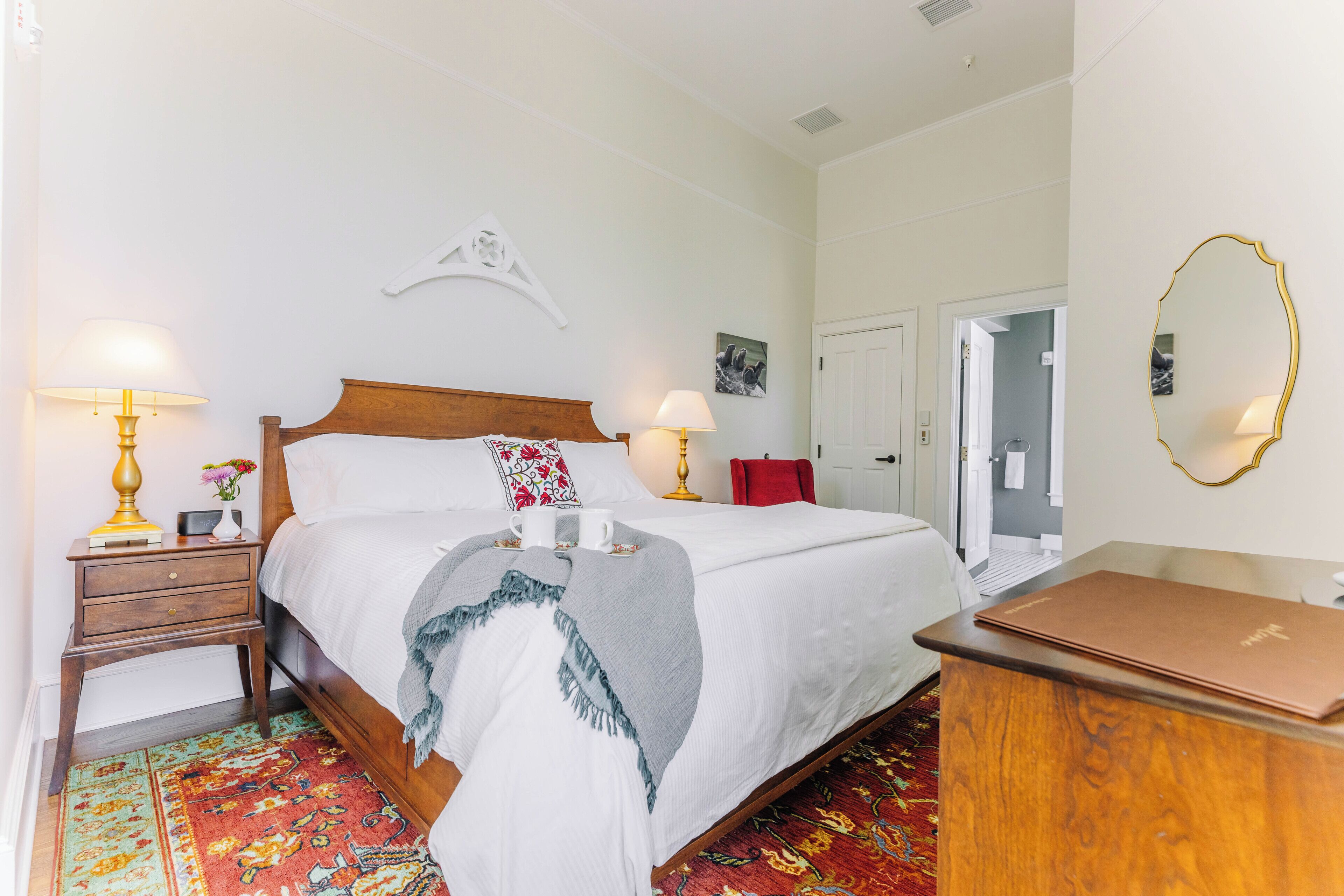 A neatly arranged bedroom with a bed, bedside lamps, a bedside table with a clock, a red chair, and a mirror on the wall.