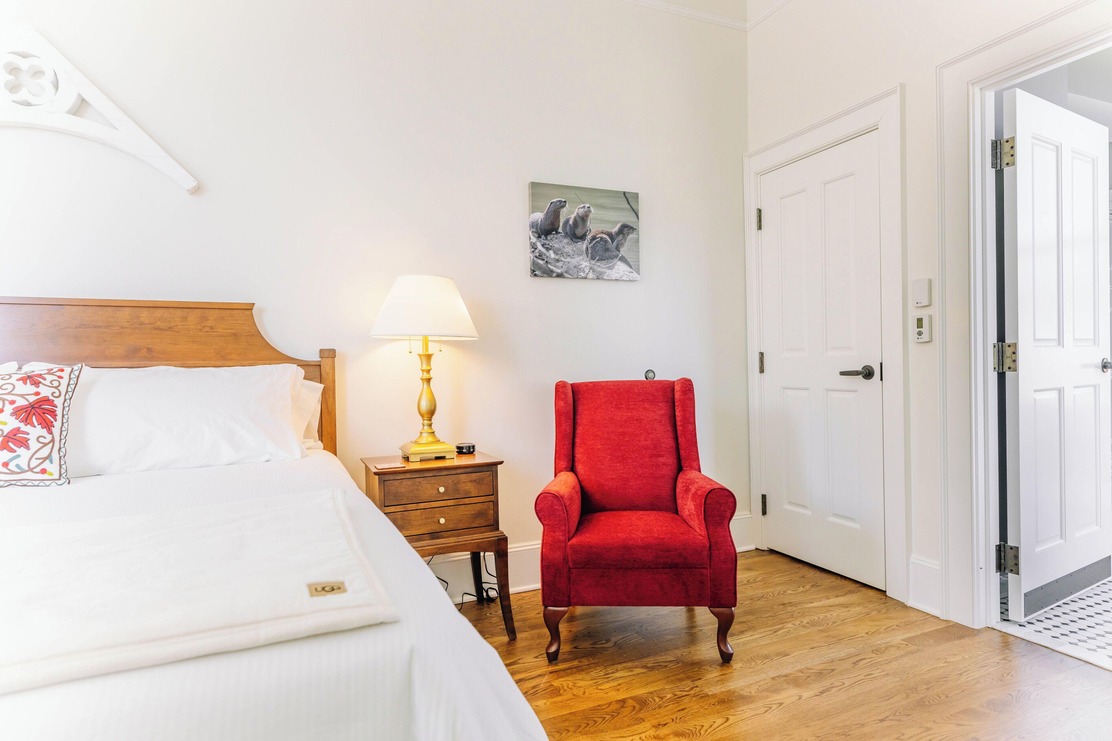A hotel room with a bed, a red armchair, a bedside table with a lamp, a picture on the wall, and a door.