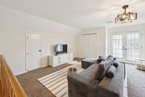 A modern living room with a sectional sofa, a flat-screen TV on a white cabinet, and a chandelier. - 5-acre Historic Home With Pool Fishing Pond Game Room Outdoor Bar (Augusta)
