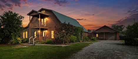 A two-story wooden house with a balcony, surrounded by greenery and a gravel driveway.