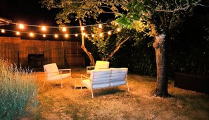 Un espacio al aire libre con dos sillas y una mesa, rodeado por un árbol y luces colgantes.
