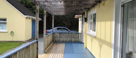 Un espace extérieur abrité doté d’une terrasse en bois, une voiture garée à l’intérieur et un bâtiment à la façade jaune.