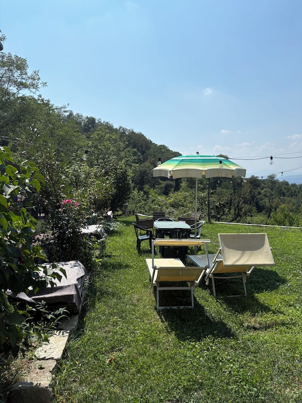 An outdoor seating area with tables and chairs, a green and white umbrella, and a view of a forested hillside.