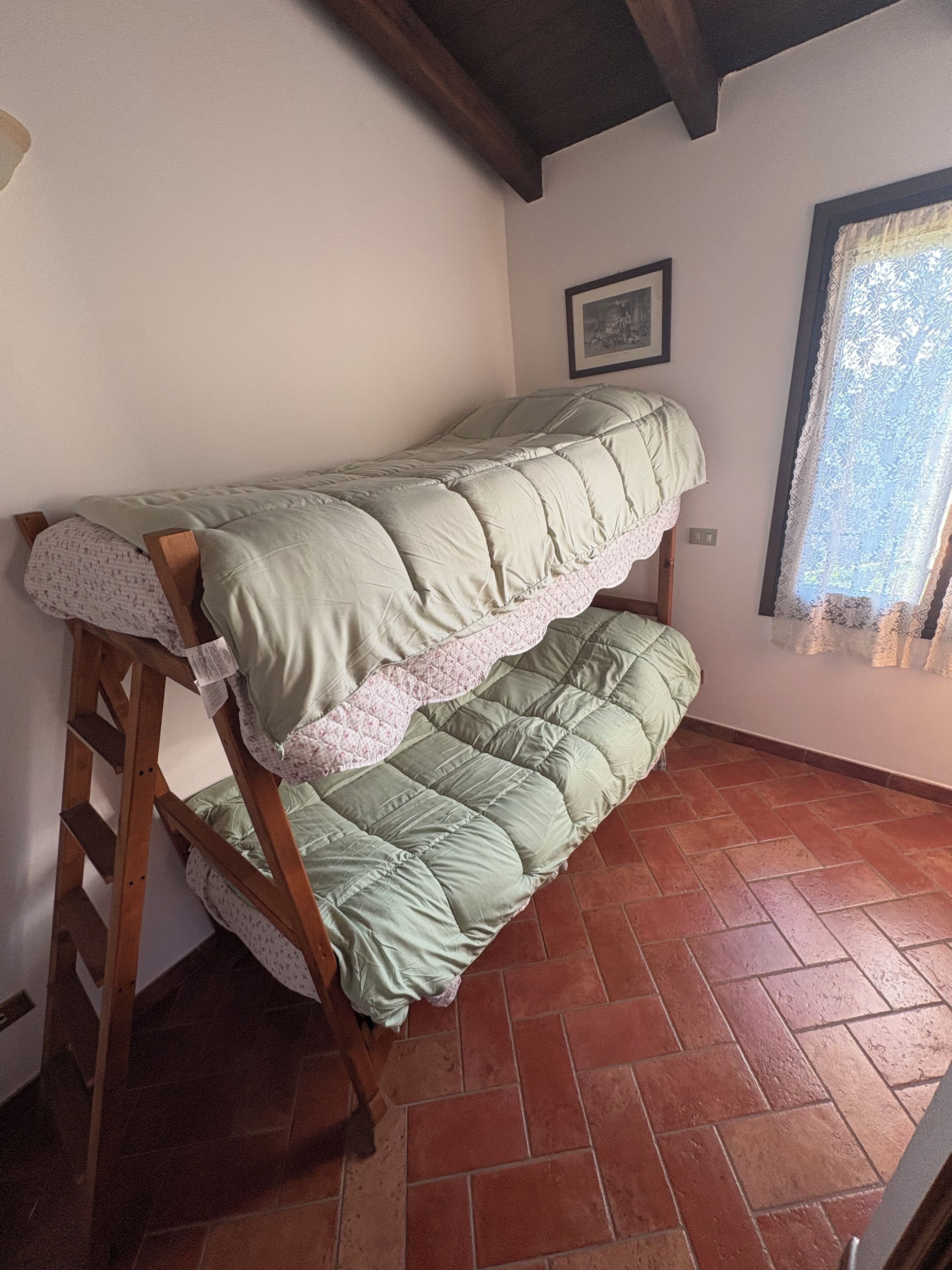 Ein Etagenbett mit einer Holzleiter, ein Fenster mit Spitzgittervorhängen und ein Fliesenboden.