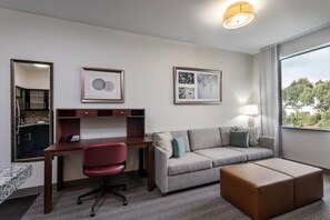 A hotel room with a desk, a red office chair, a grey sofa, a brown ottoman, and a large window with a view of trees.