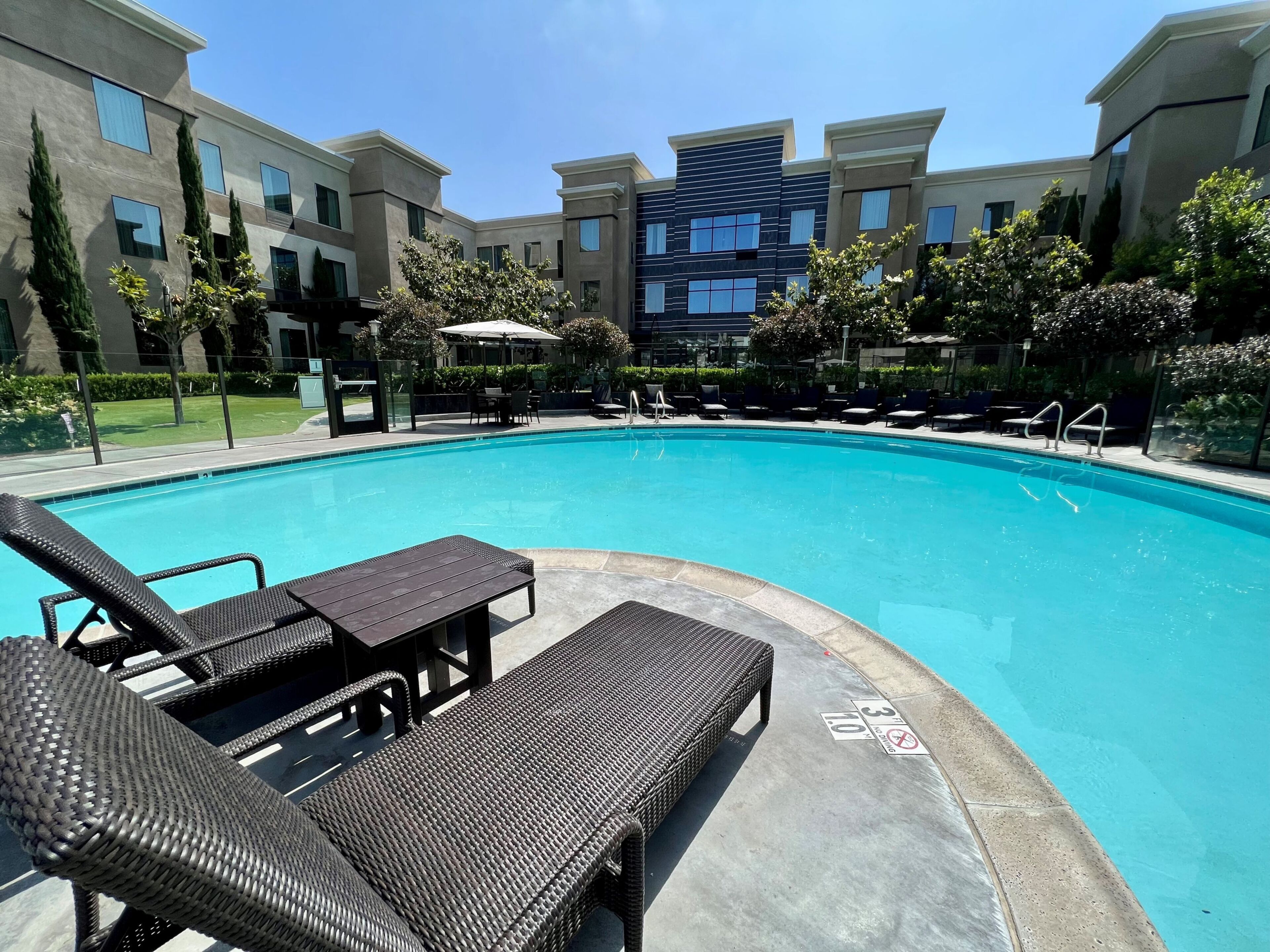 A swimming pool with lounge chairs and a table, surrounded by a landscaped area with trees and a building in the background.