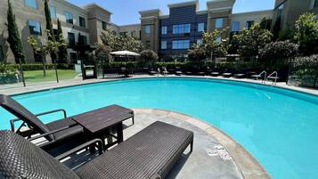A swimming pool with lounge chairs and a table, surrounded by a landscaped area with trees and a building in the background.