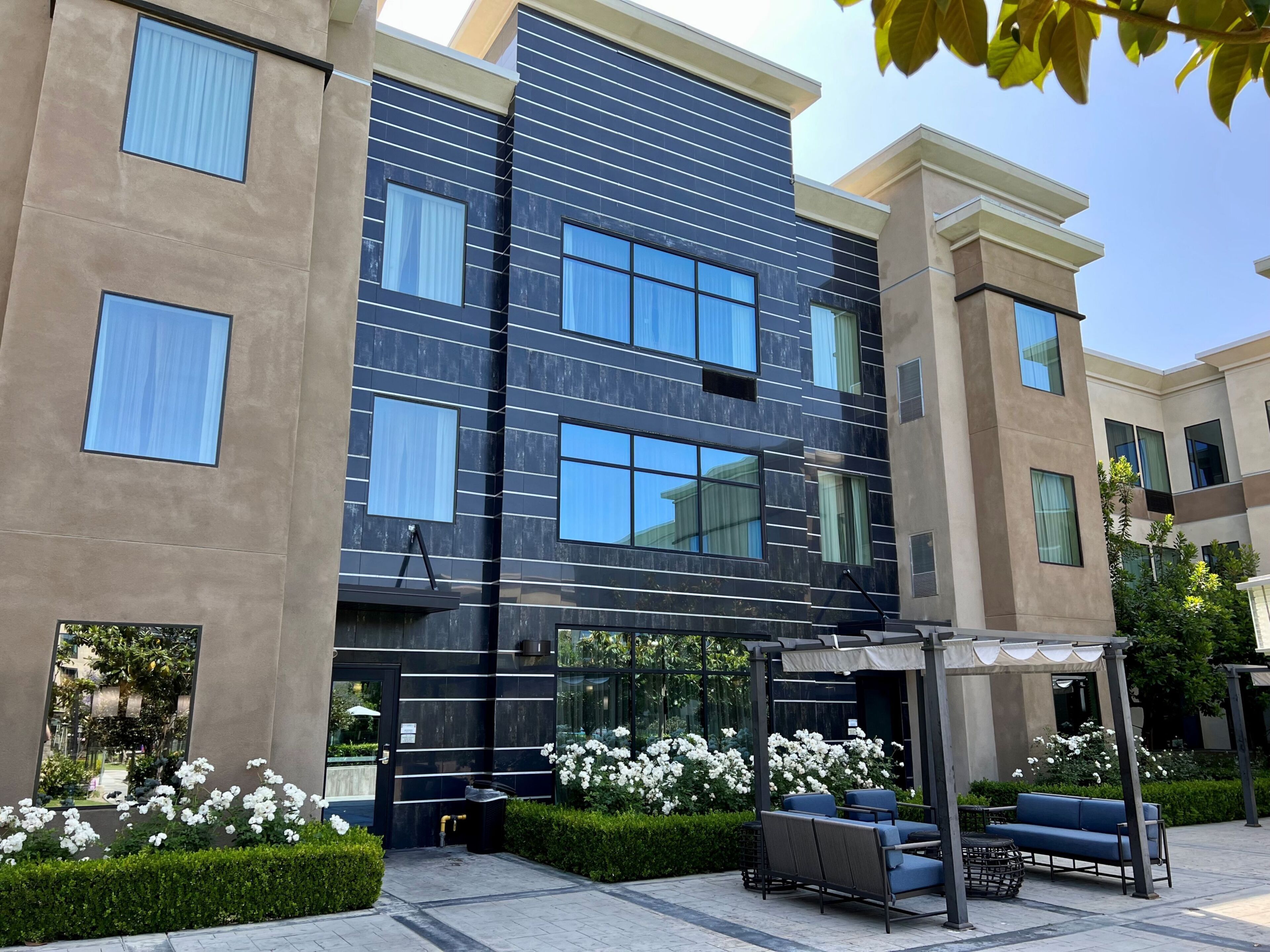 A modern building with multiple windows, a covered outdoor seating area, and a well-maintained garden with white flowers.
