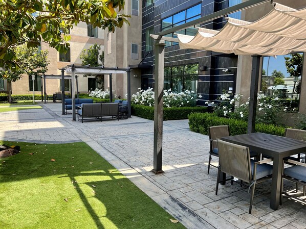 An outdoor seating area with tables and chairs, a pergola with a canopy, and a building with large windows in the background.