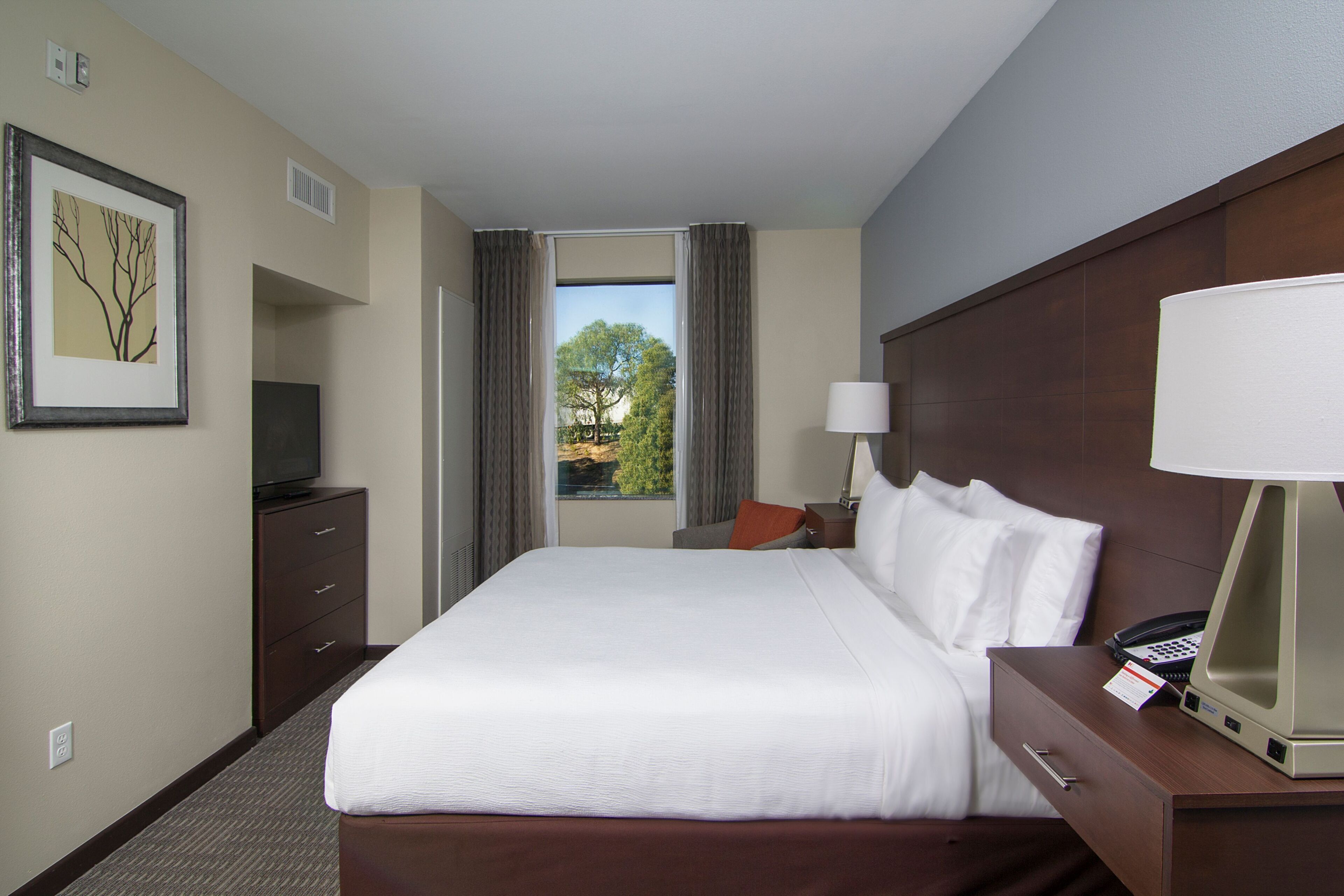 A hotel room with a large bed, a desk with a lamp, a television, and a window with curtains.