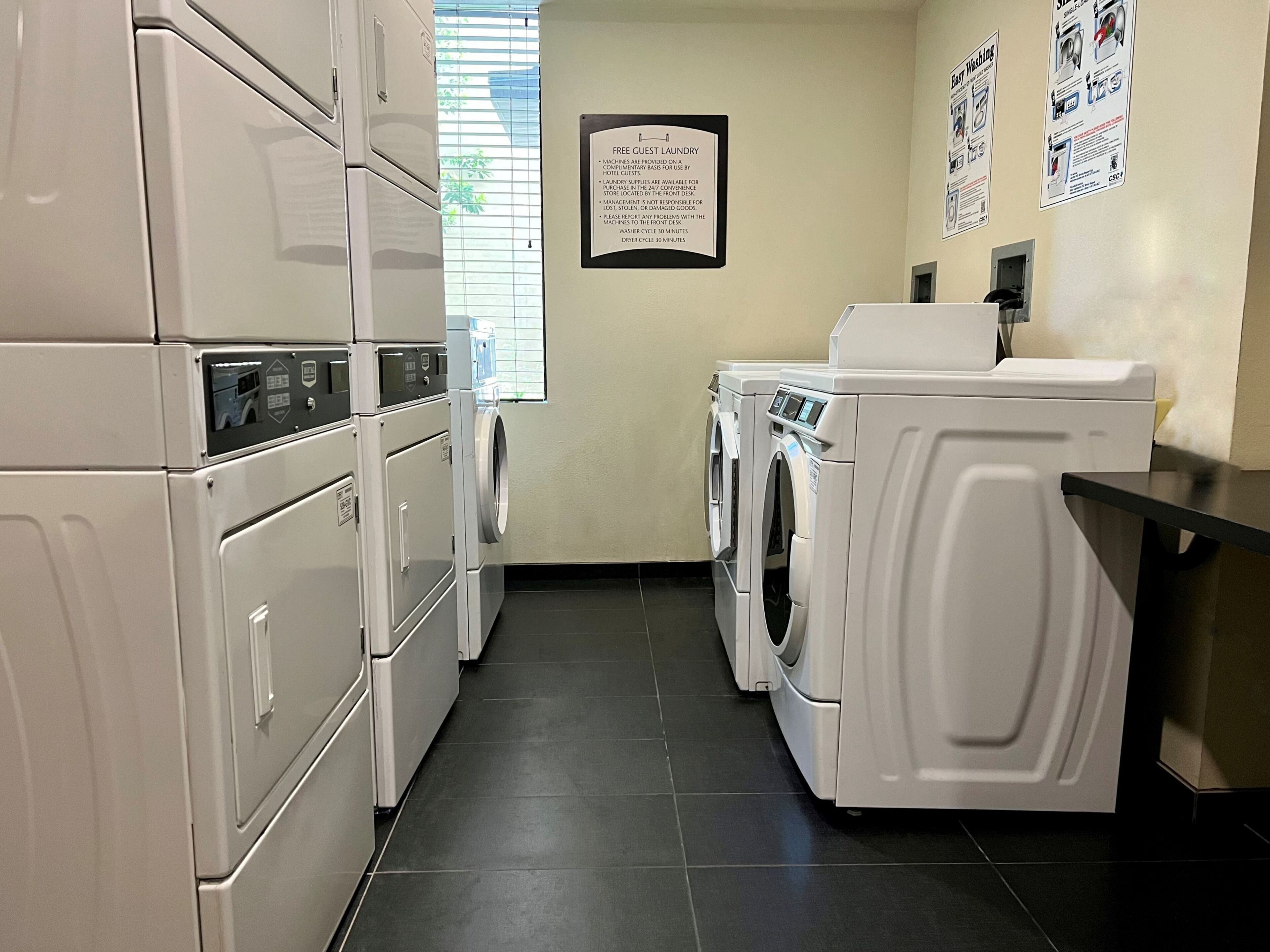 A hotel laundry room with stacked washers and dryers, a sign detailing guest laundry services, and a window with blinds.