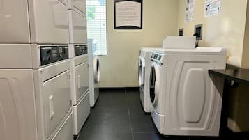 A hotel laundry room with stacked washers and dryers, a sign detailing guest laundry services, and a window with blinds.