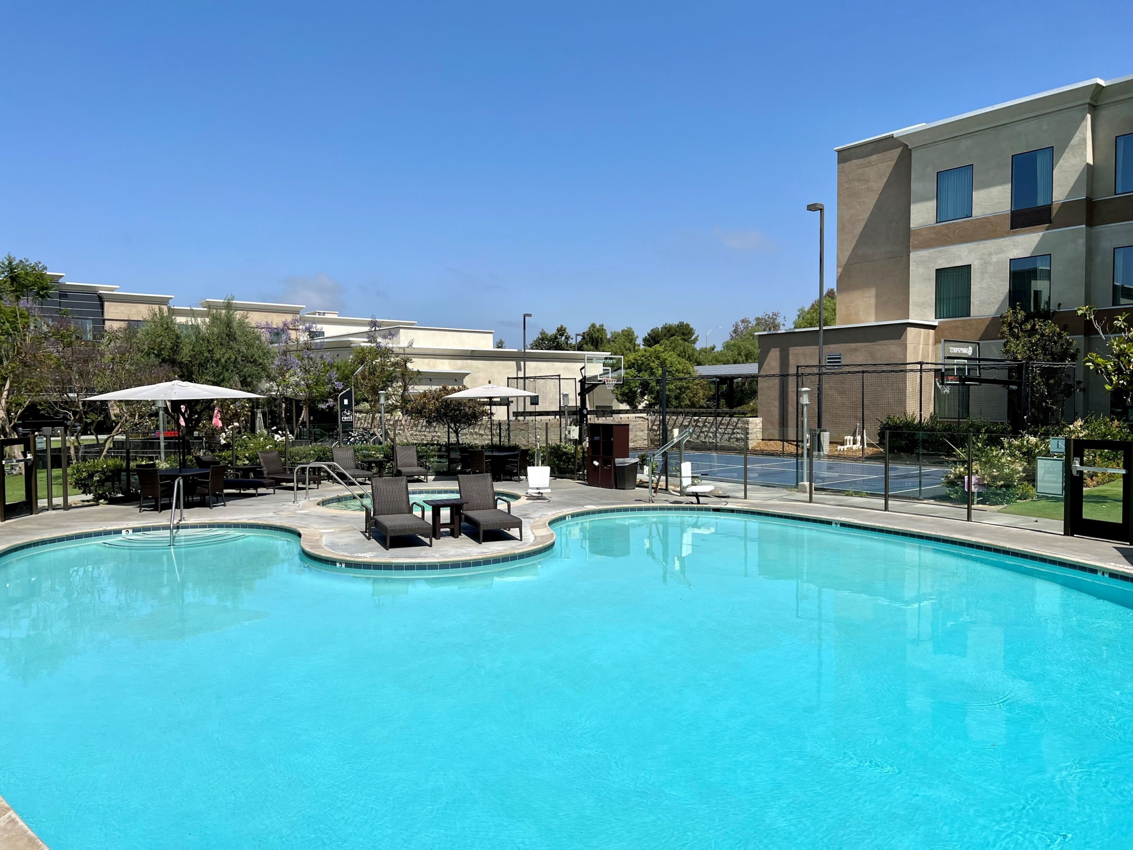 A swimming pool with lounge chairs and umbrellas, surrounded by a fence and buildings.