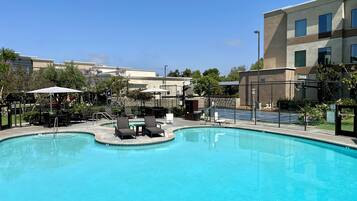 A swimming pool with lounge chairs and umbrellas, surrounded by a fence and buildings.