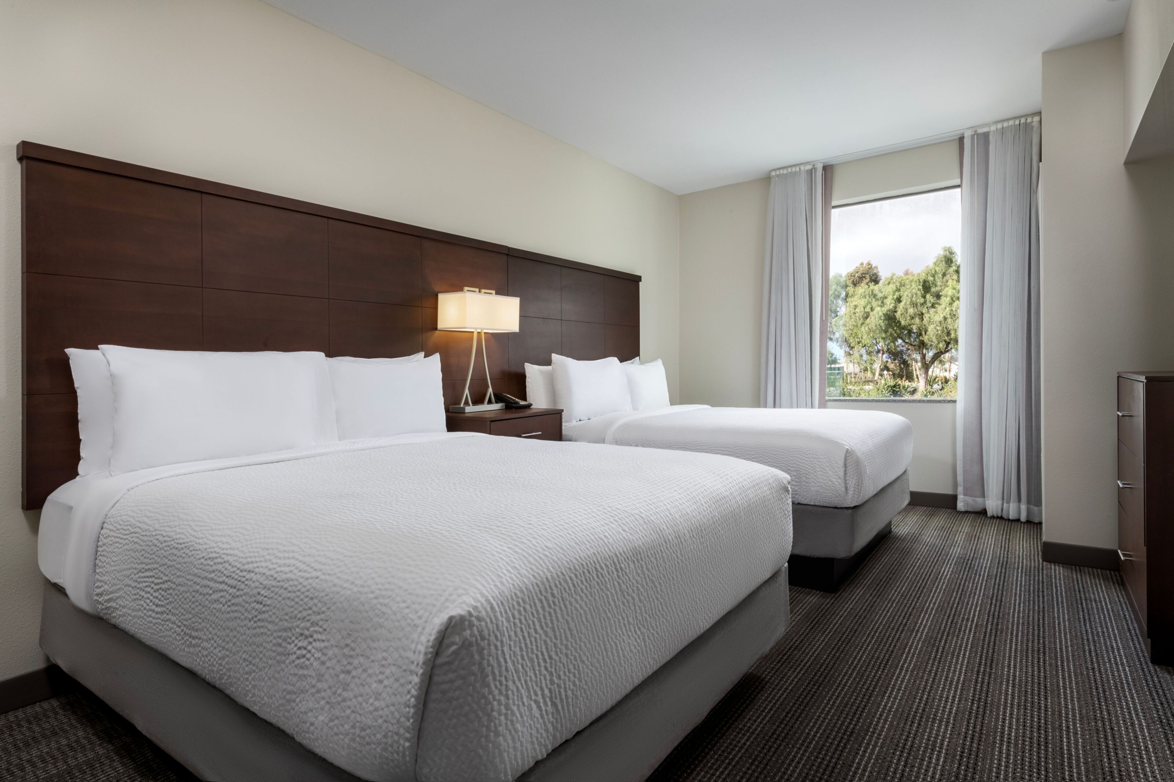 A hotel room with two beds, a nightstand, a lamp, and a window with a view of trees.