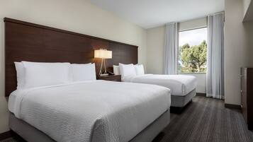 A hotel room with two beds, a nightstand, a lamp, and a window with a view of trees.