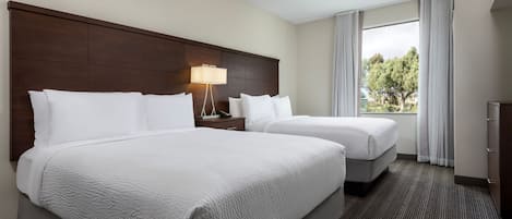 A hotel room with two beds, a nightstand, a lamp, and a window with a view of trees.