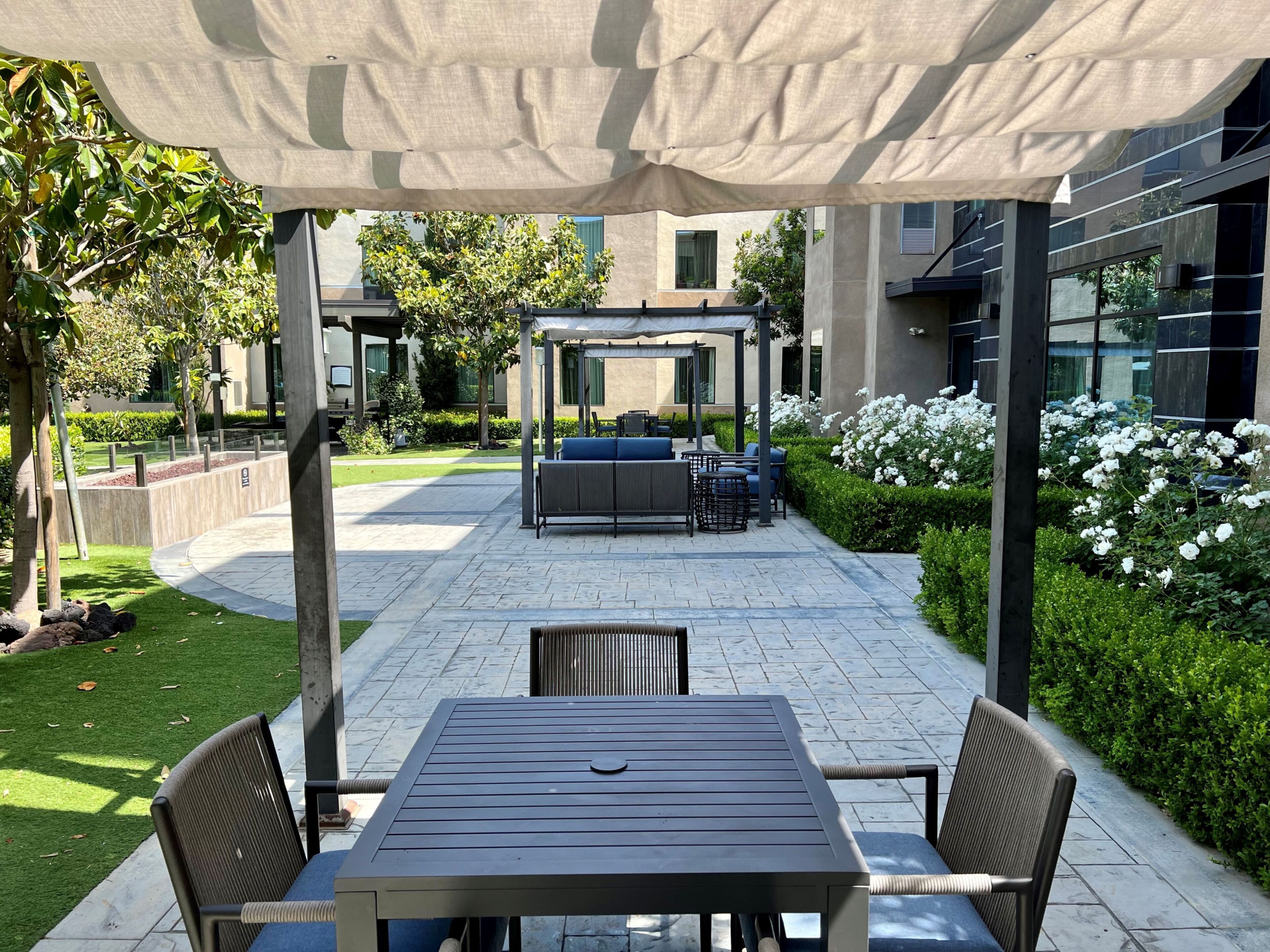 Un espace extérieur aménagé en patio, avec un coin salon couvert, une table et des chaises, et un jardin agrémenté de fleurs blanches.