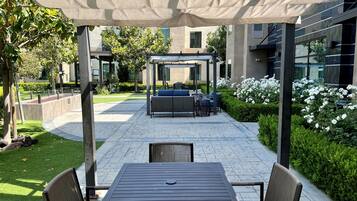 Un espace extérieur aménagé en patio, avec un coin salon couvert, une table et des chaises, et un jardin agrémenté de fleurs blanches.