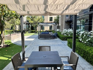 An outdoor patio area with a covered seating area, a table with chairs, and a garden with white flowers. - ADA Suite w/ Roll-in Shower | Free Breakfast (Carlsbad)