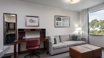 A hotel room with a desk, a red office chair, a grey sofa, a brown ottoman, and a large window with a view of trees.