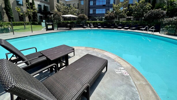 A swimming pool with lounge chairs and a table, surrounded by a landscaped area with trees and a building in the background. - ADA Suite w/ Roll-in Shower | Free Breakfast (Carlsbad)