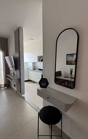 A modern interior with a white desk, a black chair, a large mirror, and a view into a kitchen area. - 2 Qtos I Praia I Lazer  I João Pessoa (Cabedelo)