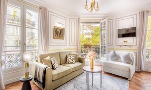 Ein gut beleuchteter Raum mit einem beigefarbenen Sofa, einem weißen Sofa, einem runden Couchtisch und einem an der Wand montierten Fernseher.