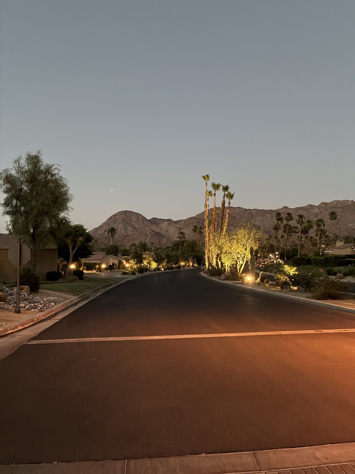 A well-lit street with a mountain in the background, surrounded by landscaped areas with trees and shrubs.