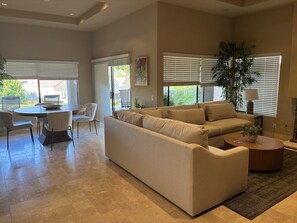 A spacious living area with a beige sofa, a dining table with chairs, and a large window with blinds. - 2.5-bedroom house in lovely Palm Desert with AC (Palm Desert)