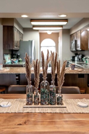 A dining table with six glass vases containing dried pampas grass and pebbles, set on a woven placemat in a modern kitchen with wooden cabinets and a large window. - Spacious Condo, Hot Tub, Sauna, Indoor  and Outdoor Pool Access! | C-11 (Branson)