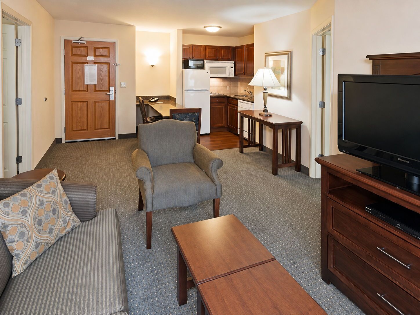 A living room with a sofa, armchair, coffee table, TV stand, and a kitchenette in the background.