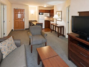 A living room with a sofa, armchair, coffee table, TV stand, and a kitchenette in the background.