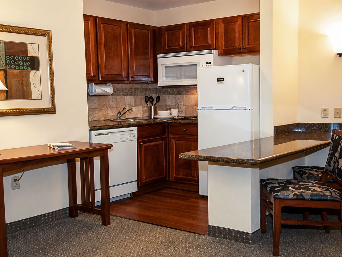A compact kitchen with wooden cabinets, a microwave, a refrigerator, a dishwasher, and a small dining area with a table and chairs.