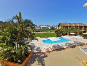Un área de piscina con sillones de descanso y sombrillas, rodeada de palmeras y un cielo azul despejado.