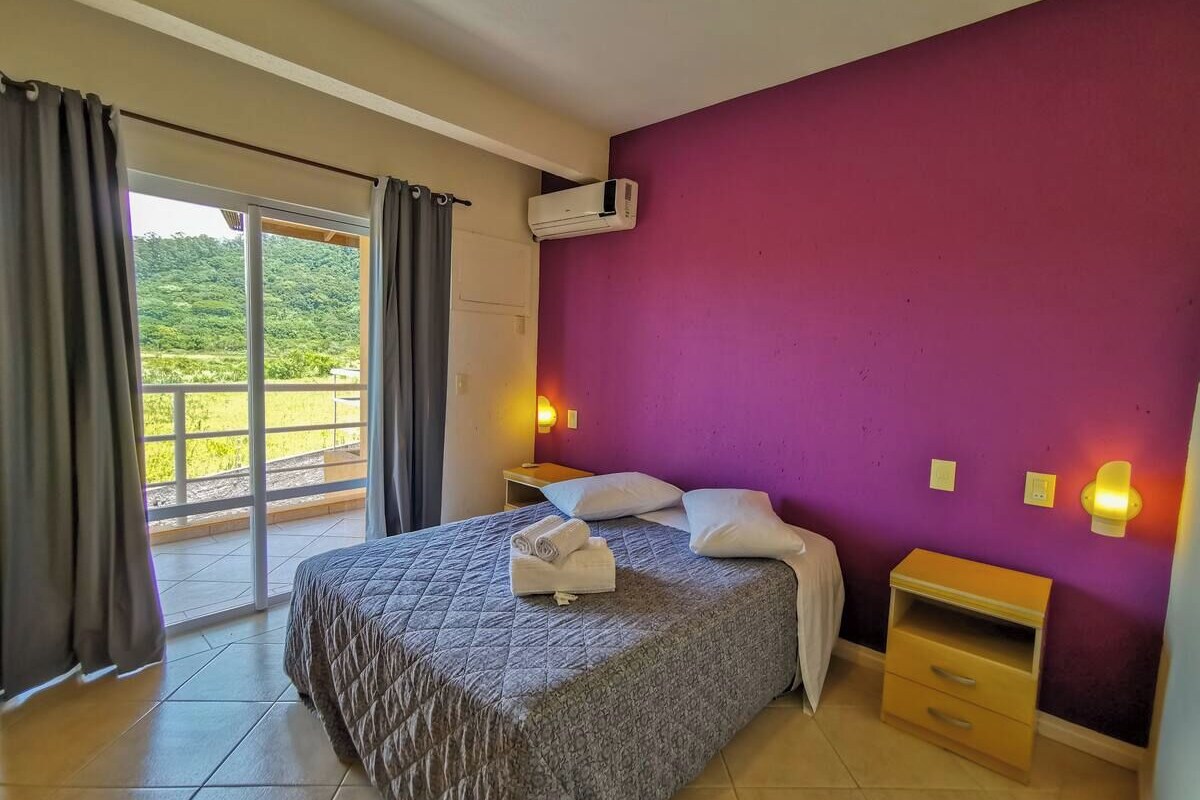 A bedroom with a bed, a nightstand, a wall-mounted air conditioner, and a view through a sliding glass door.