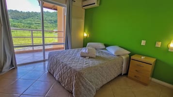 Een slaapkamer met een bed, een nachtkastje, een raam met uitzicht op groen en een airconditioning.